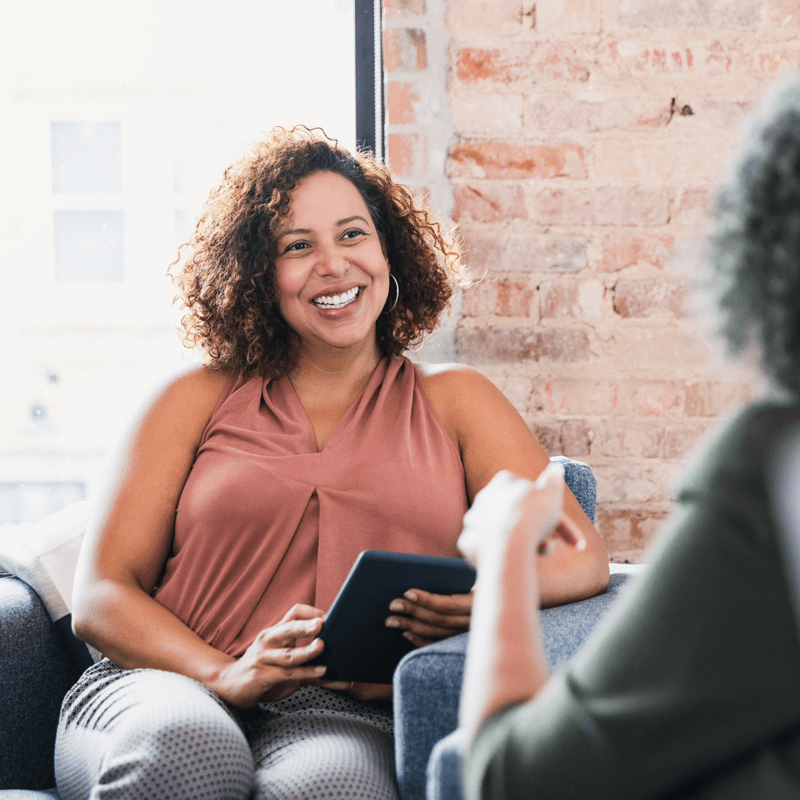 A black woman smiles holding a tablet device while seated across from a peer.